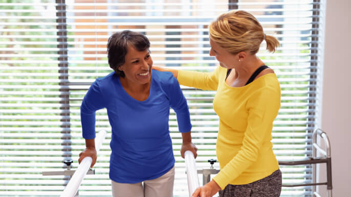 Older woman walks with support from handrails and her physical therapist.